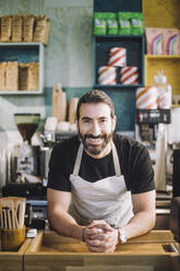 Porträt eines lächelnden männlichen Einzelhandelsangestellten, der sich mit verschränkten Händen an die Kasse eines Lebensmittelladens lehnt - MASF38648