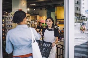 Smiling sales clerk giving shopping basket to female customer arriving at convenience store - MASF38652