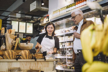 Saleswoman discussing over bread with senior male customer in grocery store - MASF38655