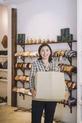 Porträt einer lächelnden Verkäuferin, die einen Karton an der Türklinke einer Modeboutique trägt - MASF38682