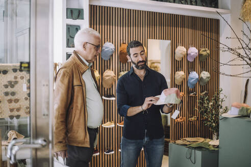 Salesman showing cap to senior male customer at clothing store - MASF38694