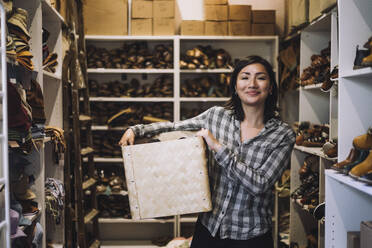 Portrait of smiling female clerk carrying wicker basket while standing amidst shoe racks at store - MASF38702