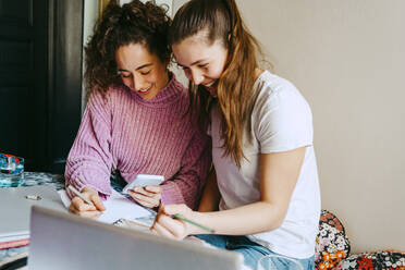 Happy young female friends using smart phone while homework - MASF38722