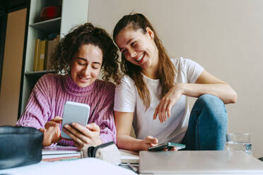 Smiling young women sharing smart phone while sitting with books at home - MASF38723