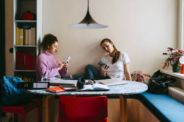 Young women using smart phones while sitting with books at home - MASF38724