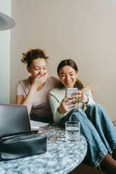 Happy young woman sharing smart phone with female teenage friend at home - MASF38743