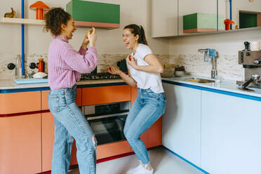 Happy young woman taking video of female friend dancing and enjoying in kitchen at home - MASF38756
