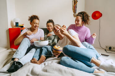 Cheerful female friends watching TV while enjoying snacks at home - MASF38761