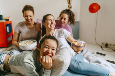 Cheerful female friends watching TV while enjoying snacks at home - MASF38764