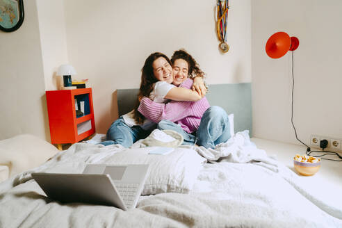 Happy young female friends embracing while sitting at home - MASF38773