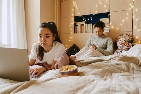 Young woman using laptop while friend using smart phone on bed at home - MASF38783