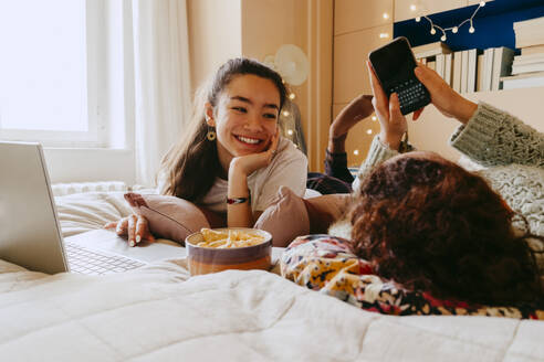 Smiling young woman using laptop while friend text messaging through smart phone on bed - MASF38787