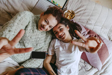 Porträt von glücklichen Freundinnen, die Friedenszeichen zeigen, während sie zu Hause auf dem Bett liegen - MASF38790