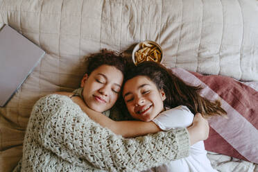 Smiling female friends embracing while lying on bed at home - MASF38791