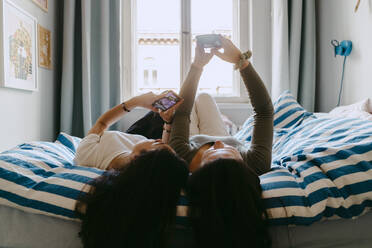 Female friends using smart phone while lying on bed at home - MASF38798