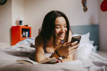 Happy young woman using smart phone while lying on bed at home - MASF38810