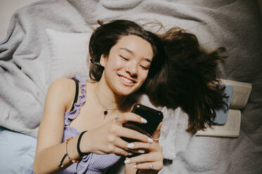 Directly above shot of smiling young woman using smart phone while lying on bed at home - MASF38814