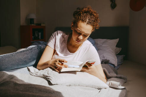 Teenage girl reading book while lying on bed at home - MASF38815