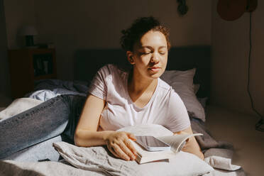 Teenage girl enjoying sunlight while while lying on bed with book at home - MASF38816