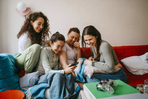 Happy young female friends looking in smart phone while sitting on sofa at home - MASF38836