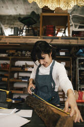 Smiling female entrepreneur measuring wood in upcycling workshop - MASF38880