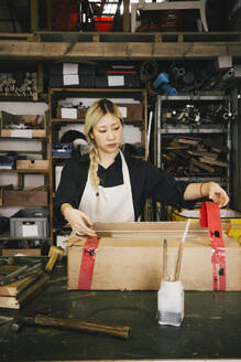 Young female entrepreneur packing cardboard box at workbench in workshop - MASF38903