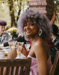 Side view portrait of smiling transwoman with curly hair sitting with friends during party in back yard - MASF38938