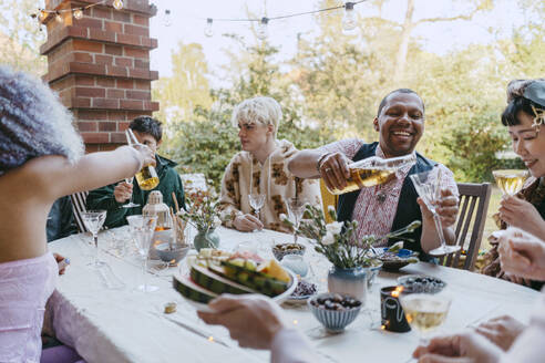 Happy friends of LGBTQ community drinking wine while sitting at table during dinner party in back yard - MASF38965