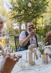 Happy gay man holding wineglass while sitting with friends during dinner party in back yard - MASF38966