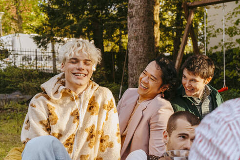 Gay friends laughing while sitting next to each other during dinner party in back yard - MASF38997