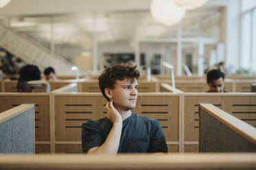 Smiling male student looking away while sitting in library at university - MASF39010