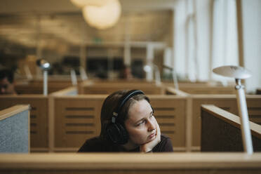 Contemplative young female student with hand on chin sitting in library at university - MASF39012