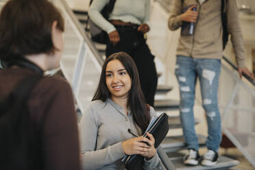 Smiling female student discussing with friend while standing by staircase in university - MASF39024