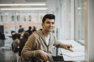 Porträt eines lächelnden männlichen Studenten, der an einem Tisch in einer Universität sitzt - MASF39036