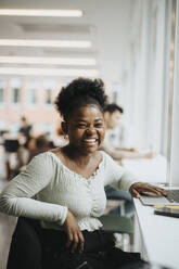 Porträt einer fröhlichen Studentin mit Laptop, die an einem Tisch in einer Universität sitzt - MASF39042