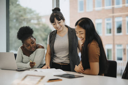 Smiling female professor explaining multiracial students at table in university - MASF39047