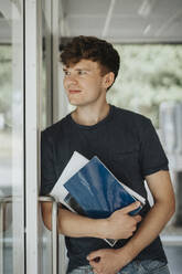 Thoughtful male holding books leaning on door in university - MASF39064