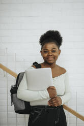 Portrait of student holding laptop standing against wall in university - MASF39065