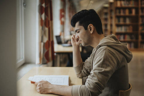 Side view of male student reading book at table in library - MASF39082