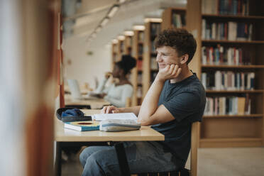 Side view of smiling student sitting with hand on chin in library - MASF39090