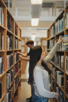 Side view of female student searching books on bookshelf in library at university - MASF39094