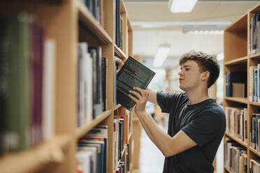 Side view of male student removing book from bookshelf in library at university - MASF39096