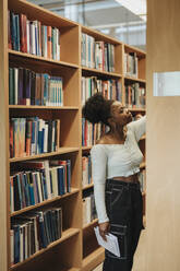 Female student searching books on shelf in library at university - MASF39098