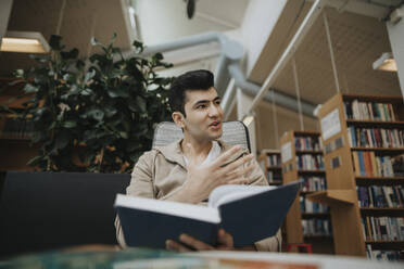 Male student gesturing while holding book in library - MASF39115