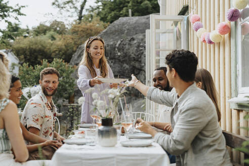 Lächelnde Frau, die einem männlichen Freund, der am Tisch sitzt, den Teller reicht, während einer Dinnerparty außerhalb eines Cafés - MASF39155
