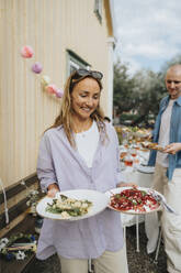 Smiling woman holding food plates while standing during dinner party at cafe - MASF39160