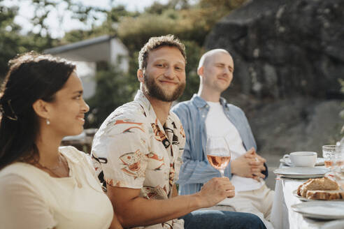 Porträt eines lächelnden jungen Mannes, der ein Weinglas in der Hand hält, während er mit Freunden beim Abendessen in einem Café sitzt - MASF39177