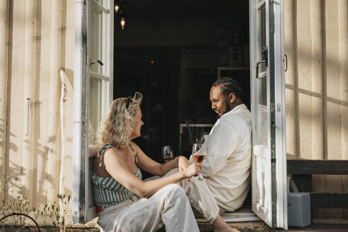Man and woman enjoying wine while talking to each other sitting at cafe doorway - MASF39186