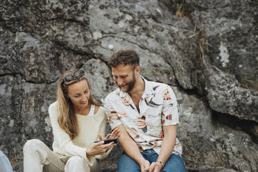 Lächelnde Frau, die ihr Smartphone mit einem männlichen Freund teilt, der vor einem Felsen sitzt - MASF39209