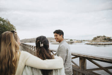 Smiling young man with female friends walking on bridge near lake - MASF39225